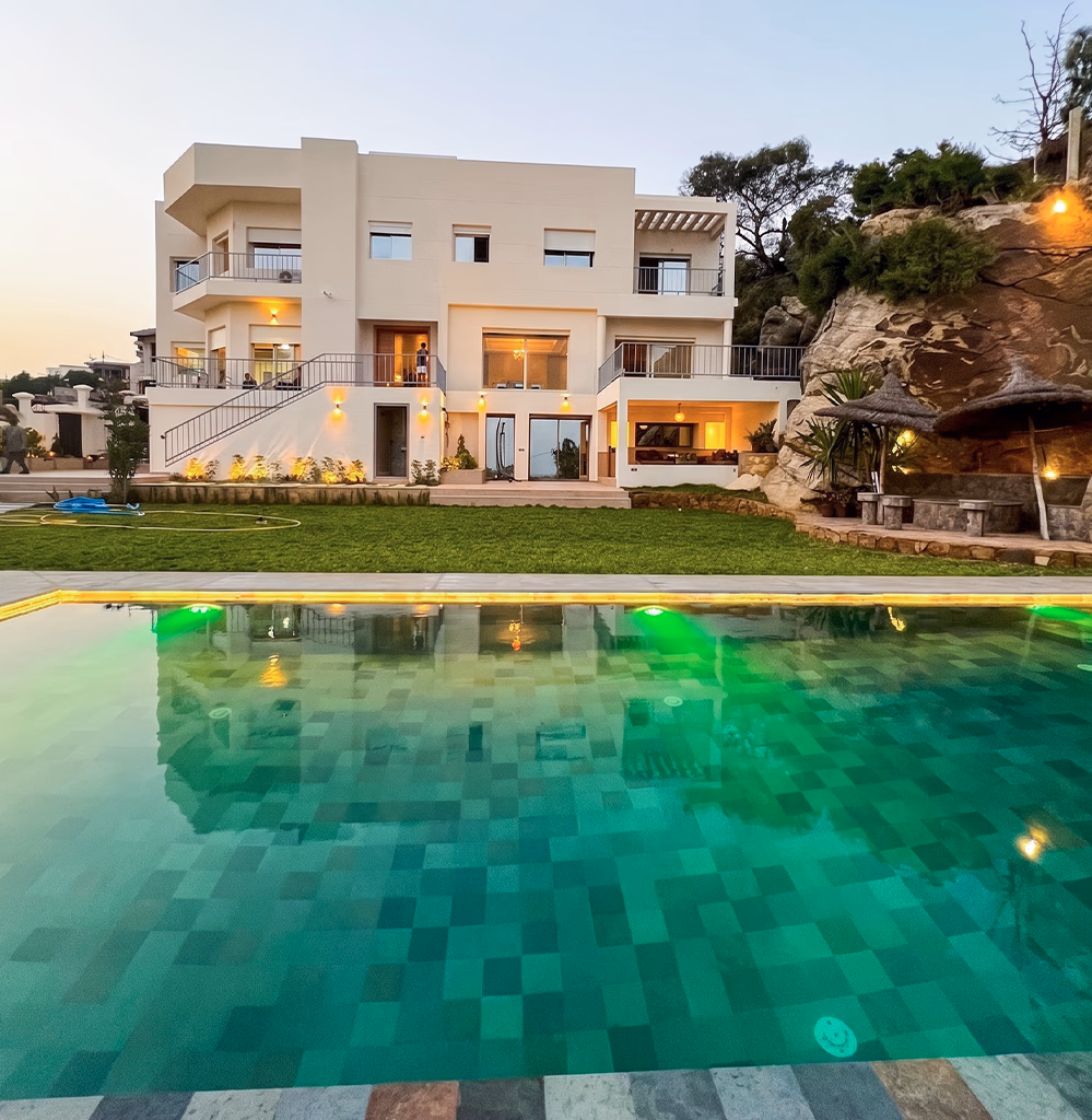 Acheter une villa à Tanger — villa contemporaine illuminée le soir avec grande piscine éclairée, balcons et jardin paysager, programme immobilier accompagné par l'agence Conciera à Achakar, Cap Spartel et Malabata Hills